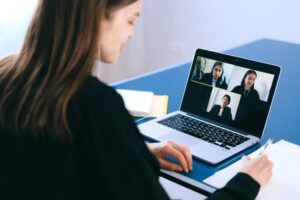 Eine Person nimmt an einer Videokonferenz mit drei weiteren Teilnehmenden über einen Laptop teil und macht sich Notizen.