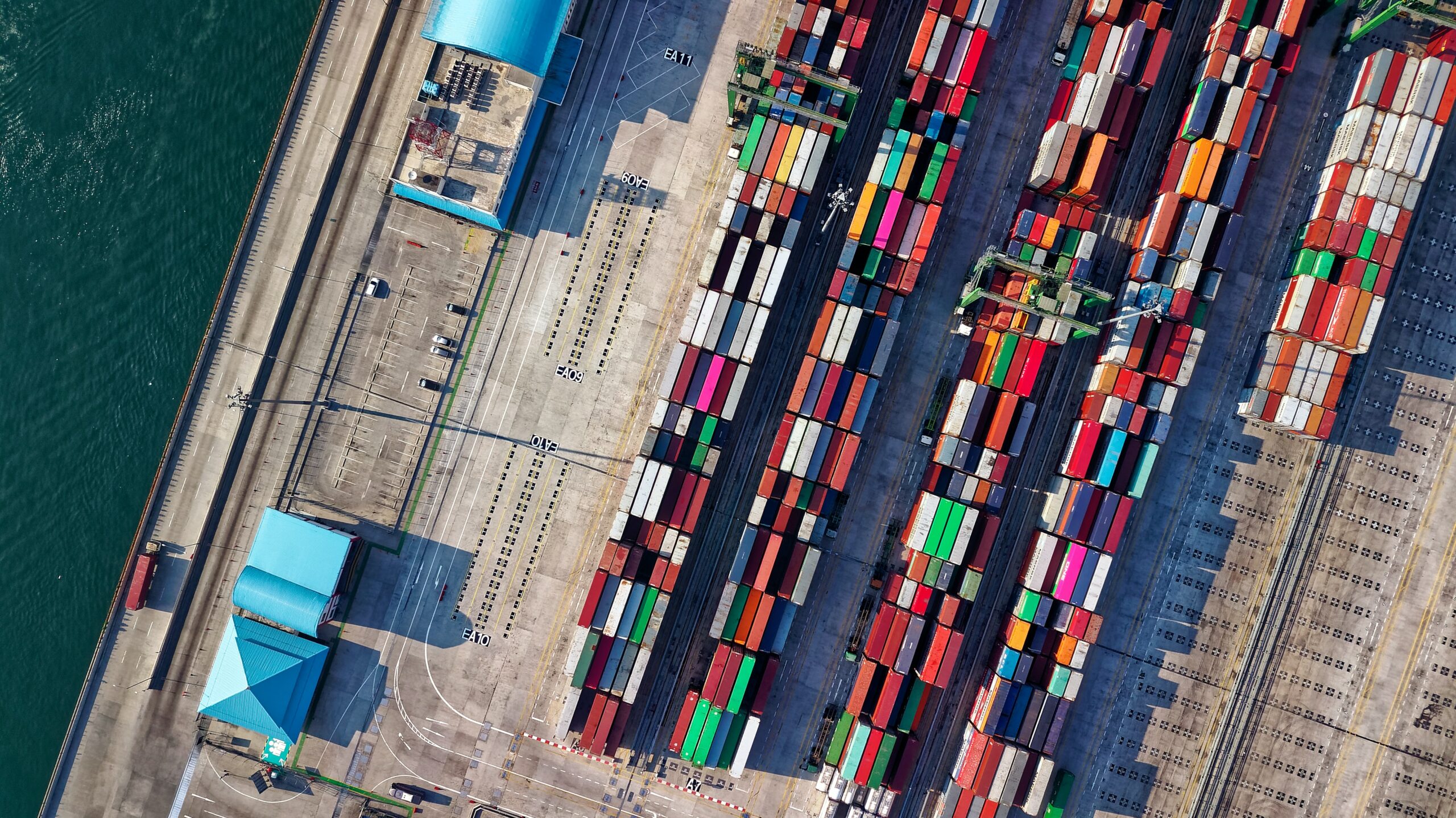 Farbig sortierte Containerreihen auf einem großen Hafengelände direkt am Wasser, Symbol für internationalen Warenverkehr.