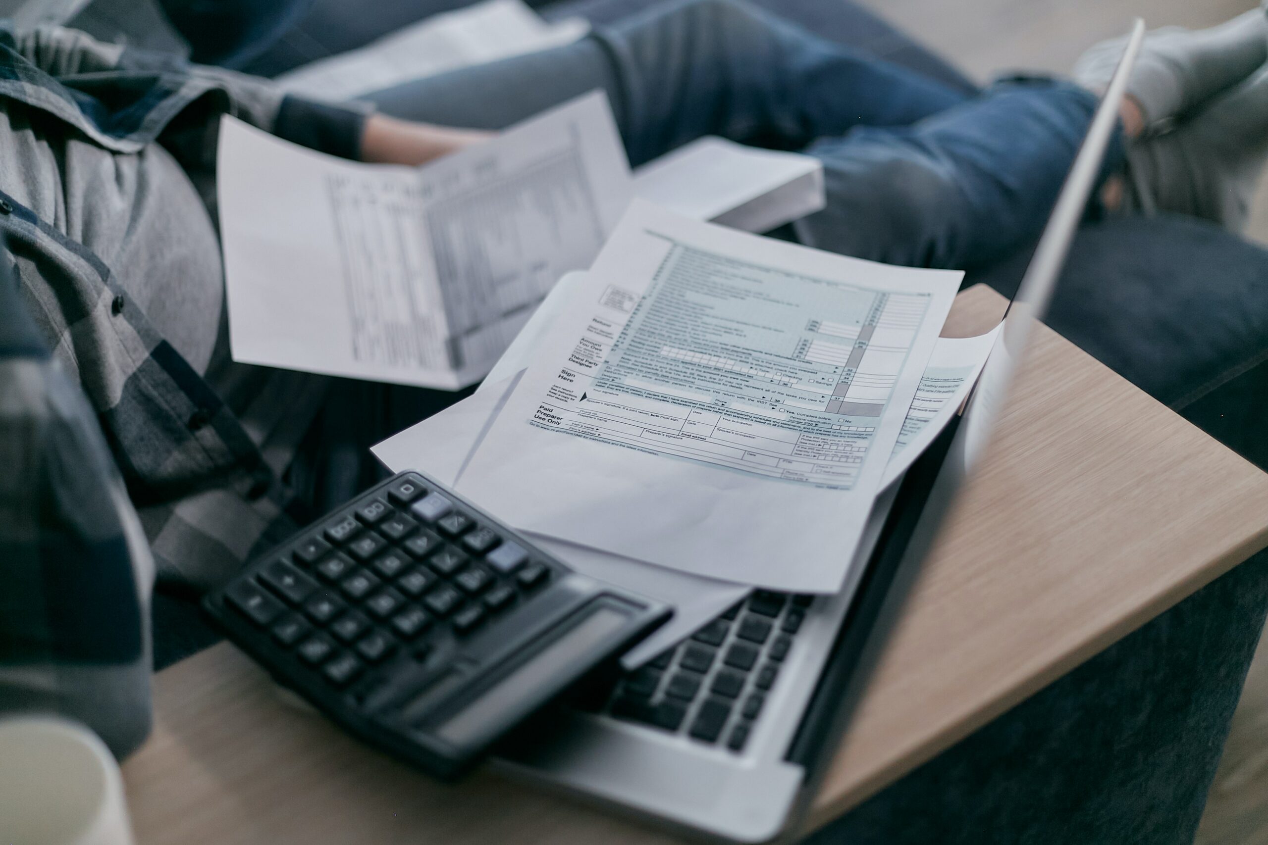 Gedruckte Formulare und ein Taschenrechner liegen auf einem Laptop, während eine Person im Hintergrund Unterlagen sortiert.