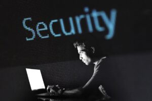 Eine Person sitzt vor einem Laptop in dunkler Umgebung, im Hintergrund ist der Schriftzug „Security“ groß eingeblendet.