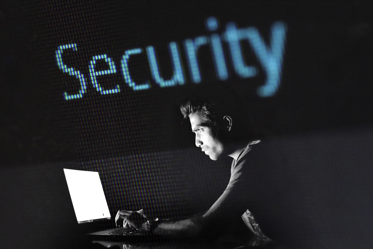 Eine Person sitzt vor einem Laptop in dunkler Umgebung, im Hintergrund ist der Schriftzug „Security“ groß eingeblendet.