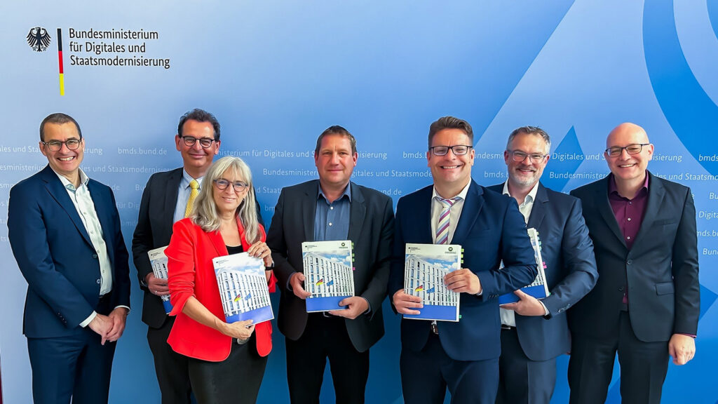 Gruppenfoto mehrerer lächelnder Personen vor einer Wand mit dem Logo des Bundesministeriums für Digitales und Staatsmodernisierung.