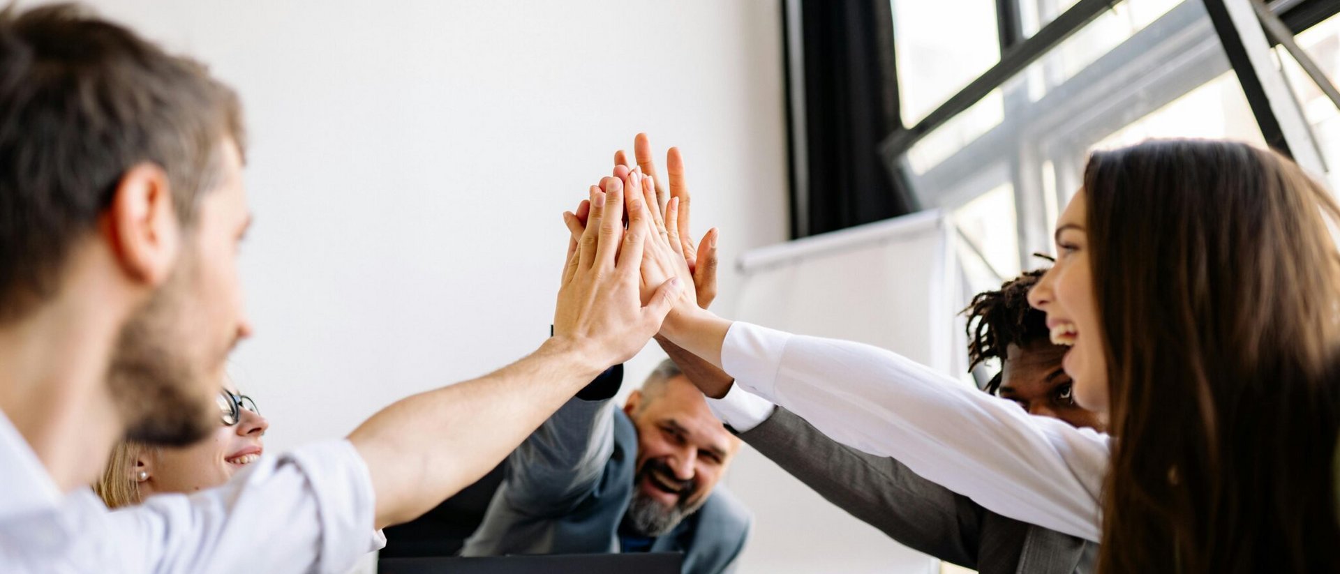 A diverse team of colleagues celebrating a success with a joint high-five in a bright office space.